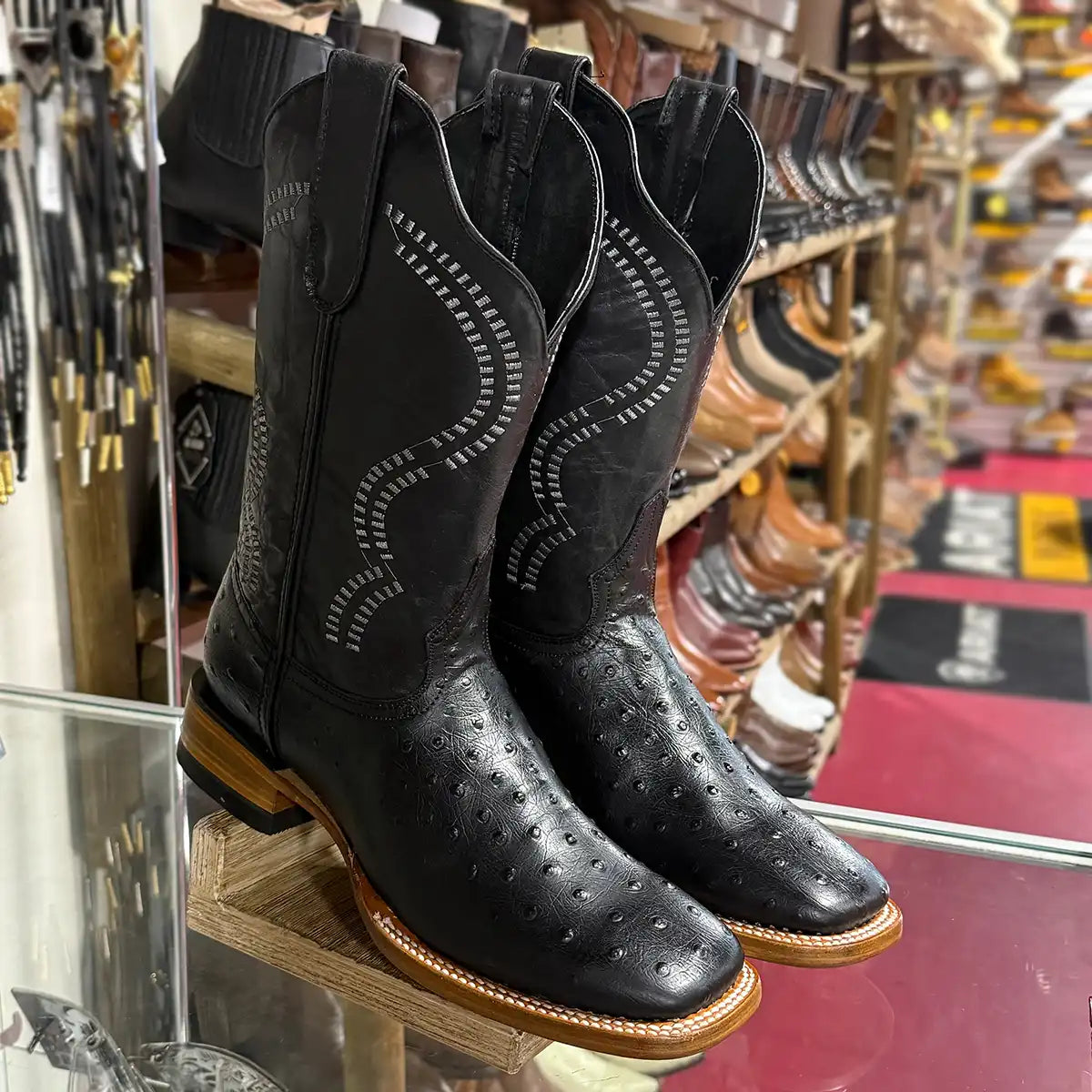 Front three-quarter angle view of White Diamonds Men's black ostrich print square toe cowboy boots displayed on a glass shelf.