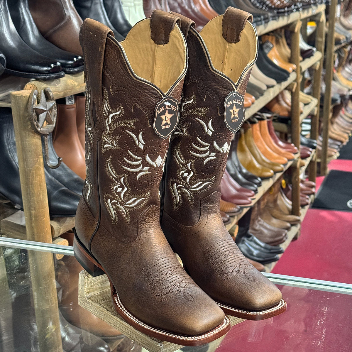 A pair of Los Altos women's brown leather square toe cowboy boots with white embroidered floral detailing and star-shaped pull tabs, displayed on a glass shelf in a western store.