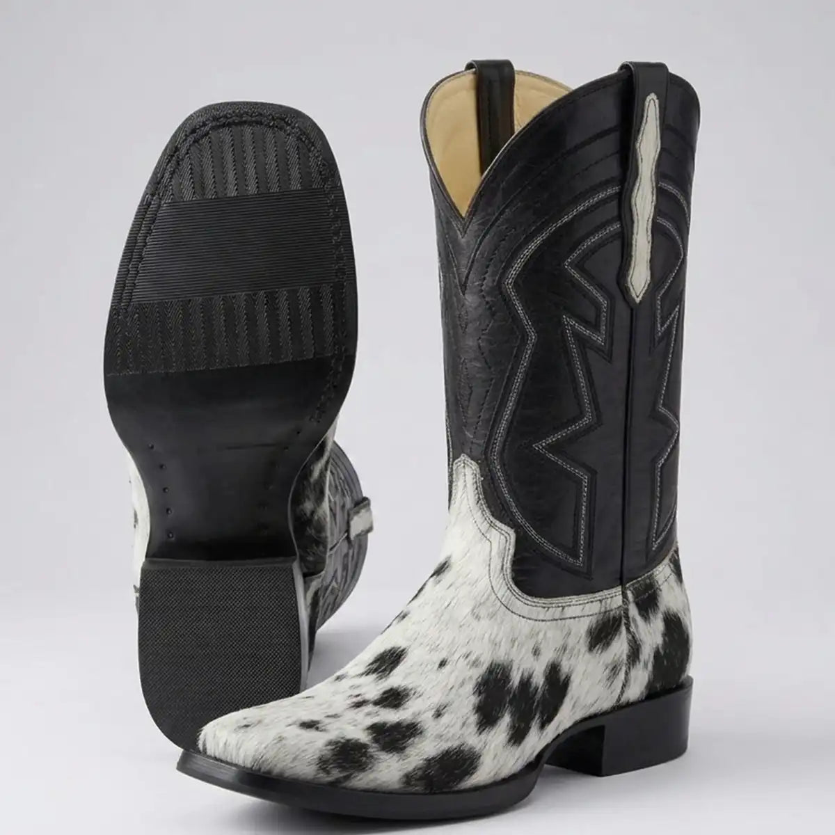 A pair of western cowboy boots shown on a light gray studio background. One boot is standing upright to show the black shaft with white stitching, black pull tab, and a black-and-white cowhide lower section.