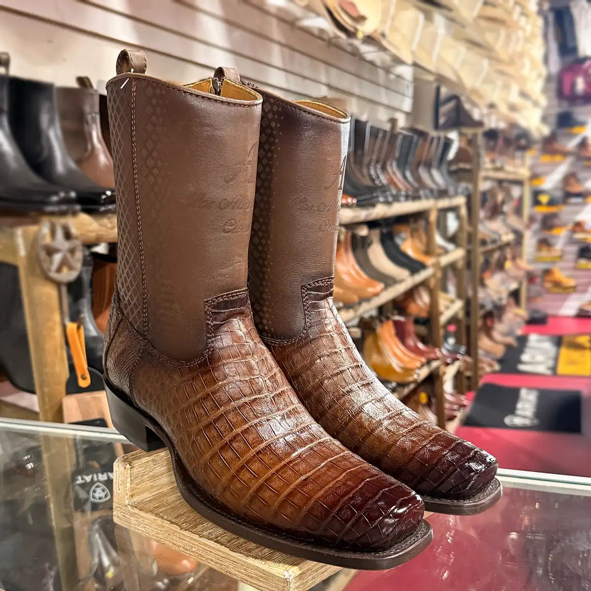 A pair of brown caiman leather cowboy boots with with a textured shaft, displayed on a wooden stand inside a boot shop.