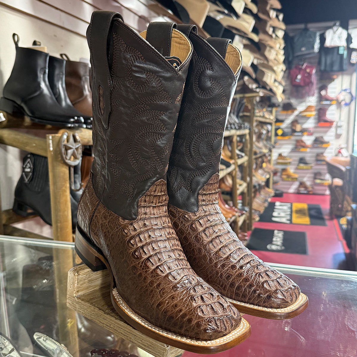 A pair of Los Altos men's brown caiman hornback leather square toe cowboy boots with a western embroidery, displayed on a wooden stand inside a boot shop.