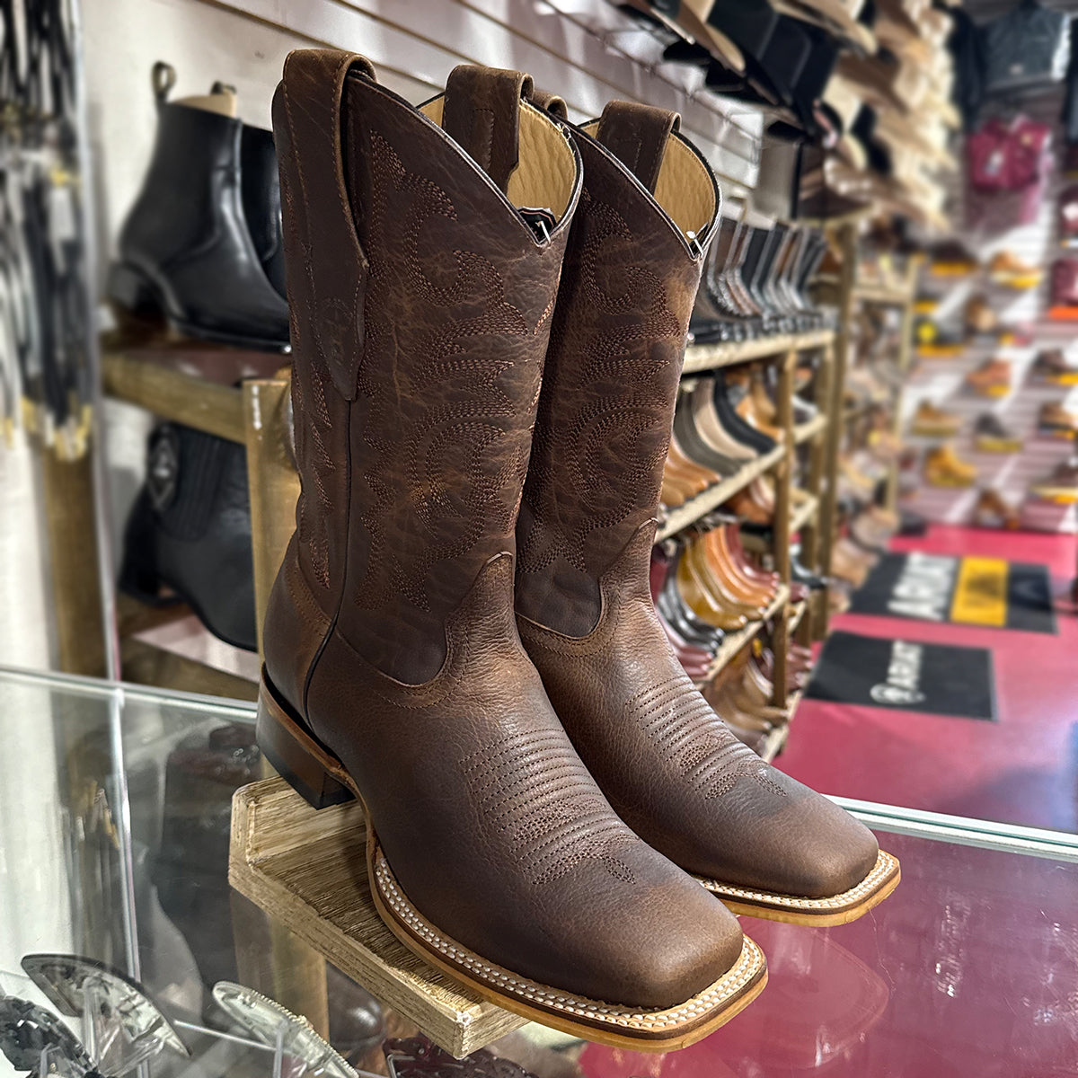 A pair of Los Altos dark brown textured leather cowboy boots with square toes, displayed on a wooden stand inside a western store.