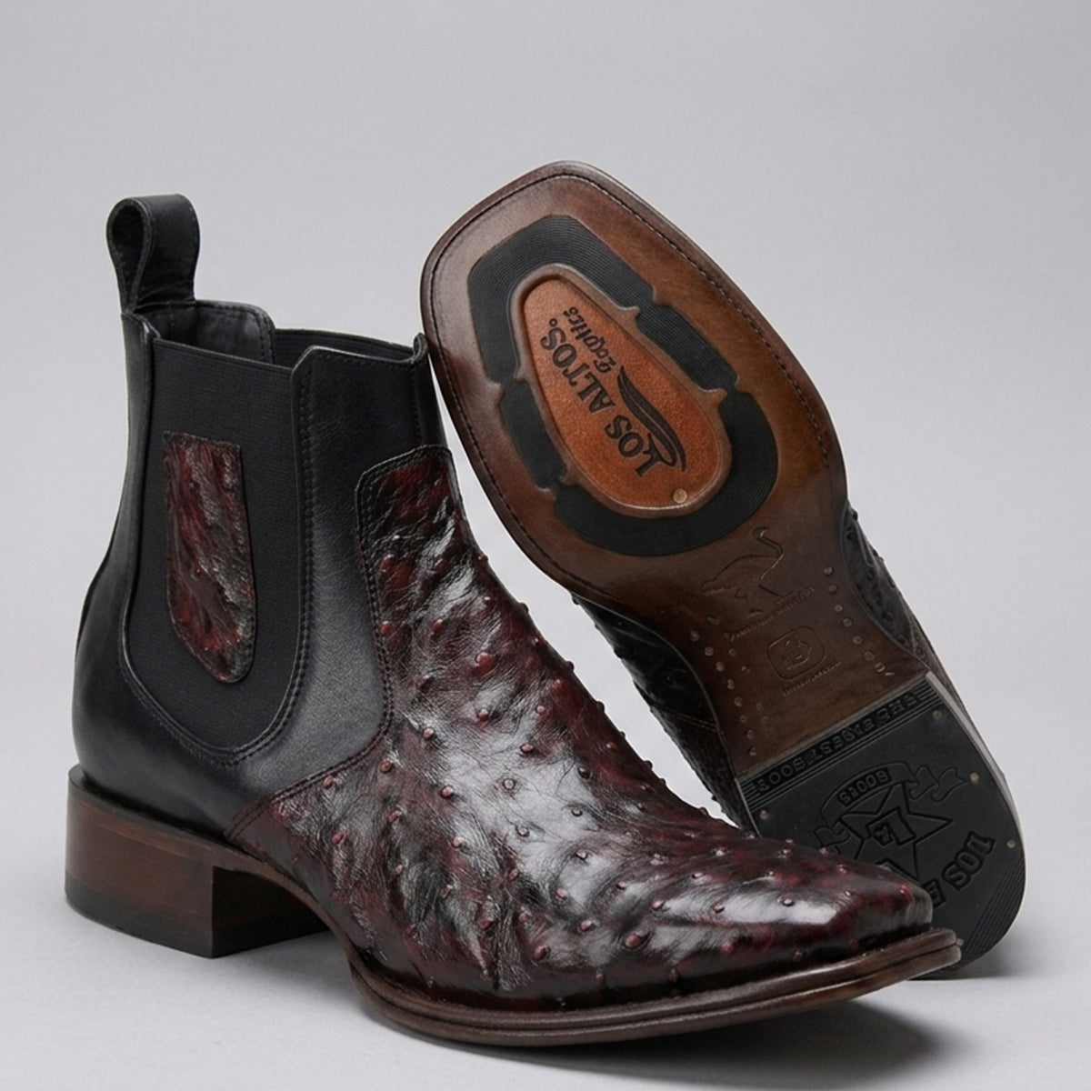 A pair of men's western ankle boots in brown and black, featuring a glossy ostrich leather vamp and black elastic side panels. One boot is shown from the side, and the other is turned to display the sole, which features a brown leather insole with 'Los Altos' branding.