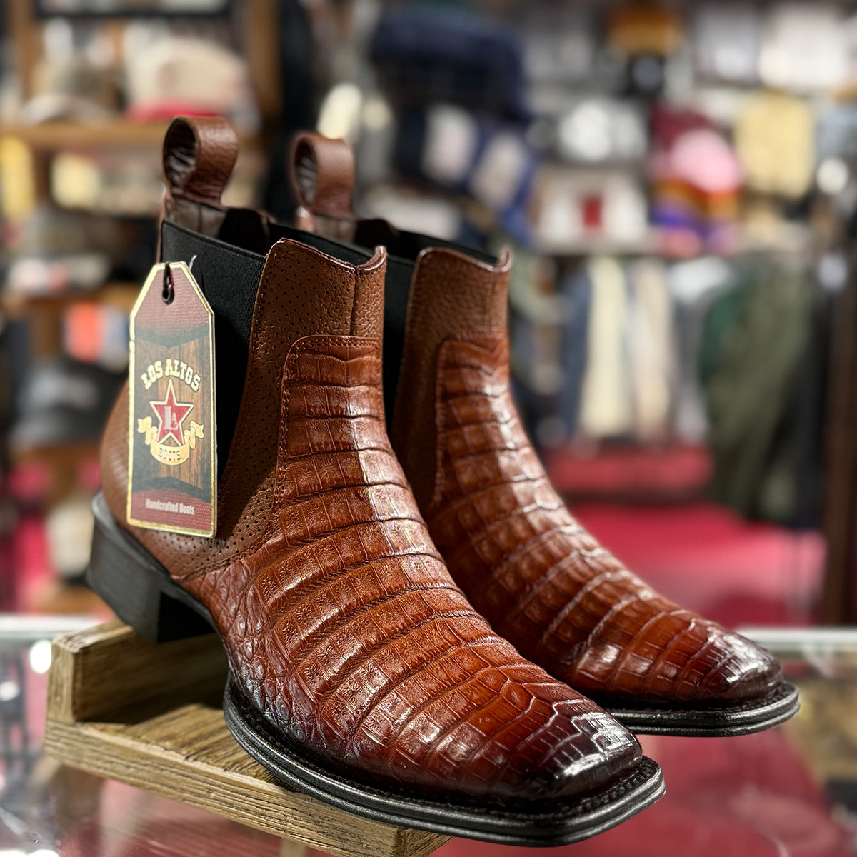 Los Altos men's brown crocodile-patterned square toe boots on a display stand with a blurred store background.