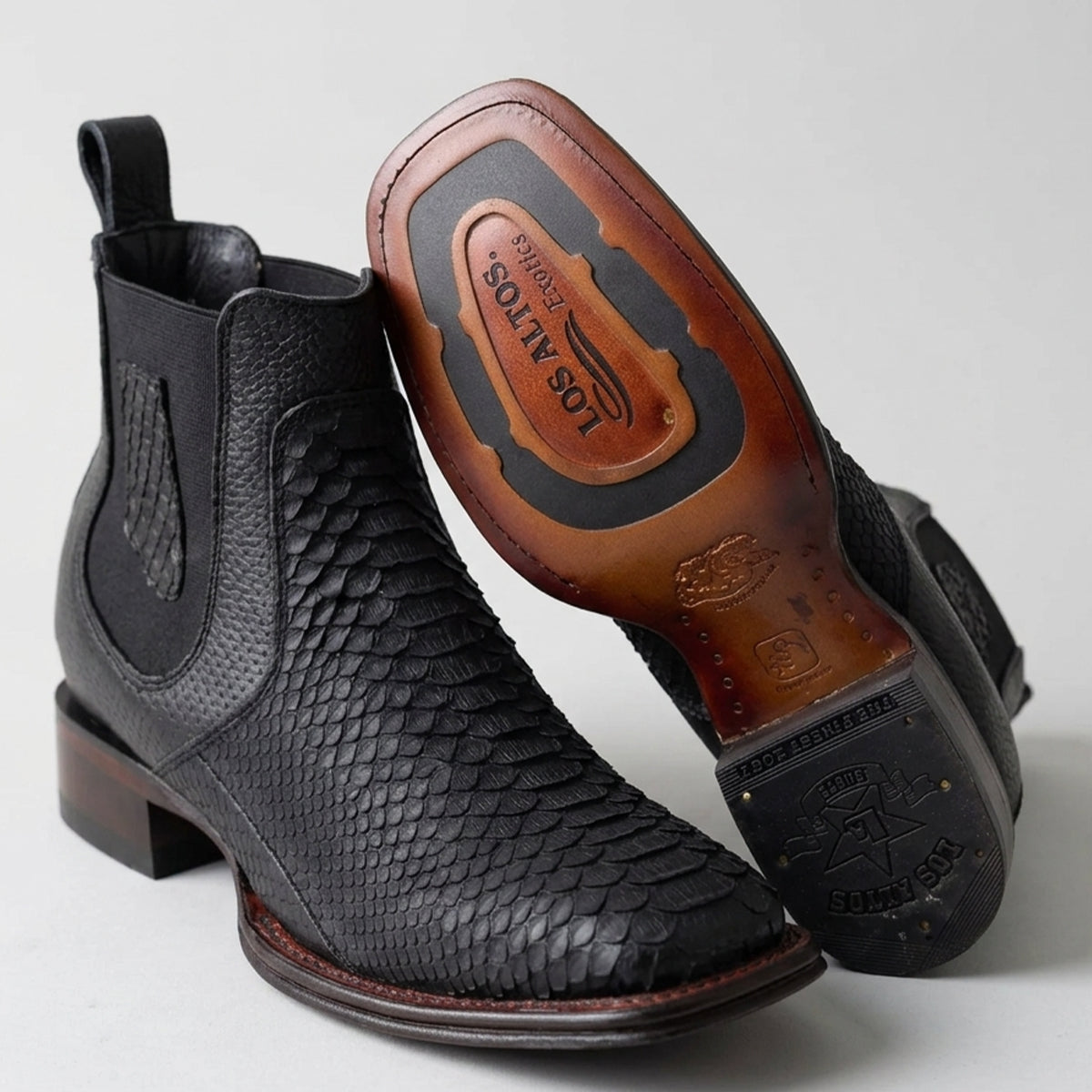 Los Altos men's black python snakeskin leather ankle cowboy boots with elastic side panels, shown on a light gray background with one boot standing upright and the other tilted to display the sole.
