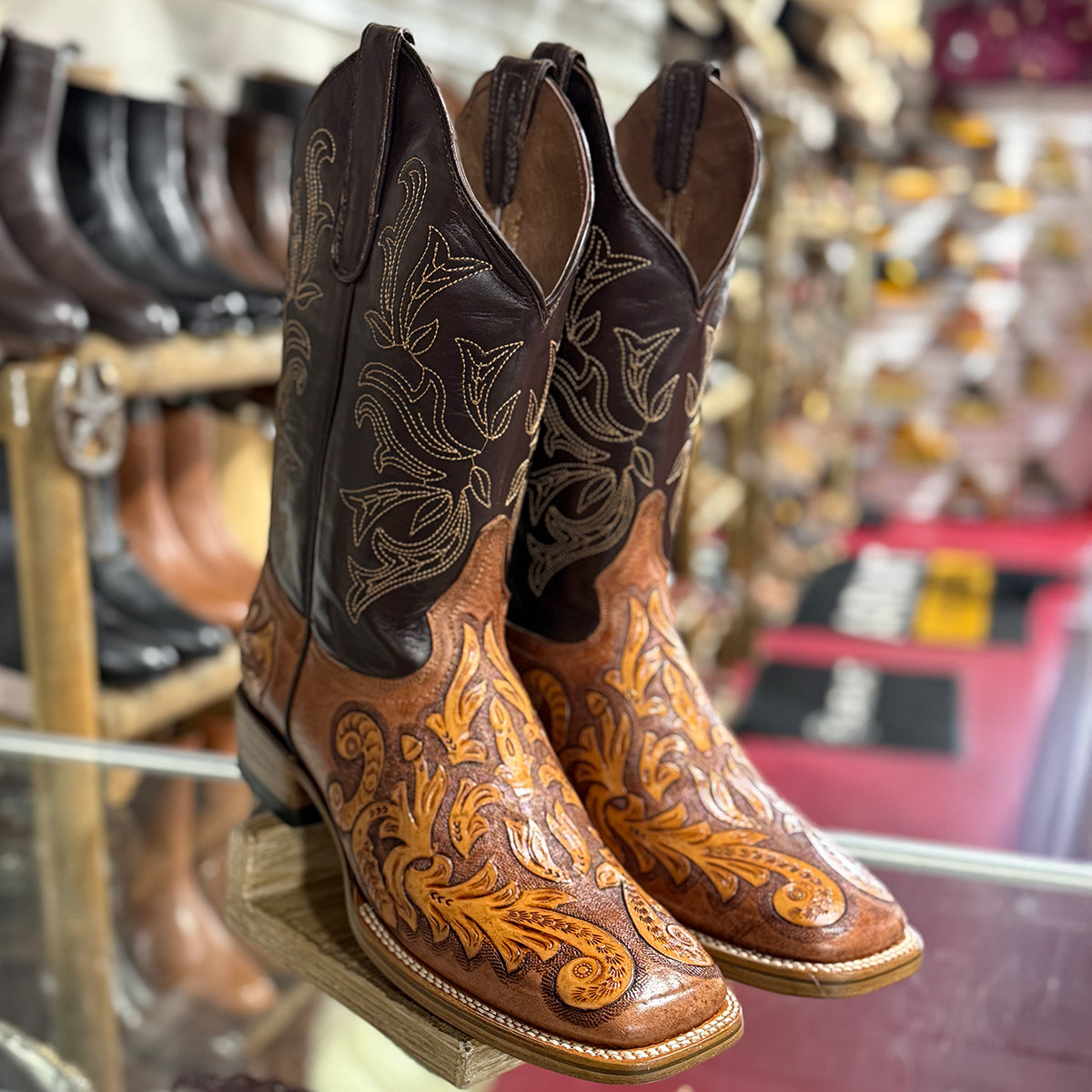 A pair of ornate western cowboy boots with dark brown shafts and tan lower shafts featuring intricate floral tooling, displayed on a glass shelf in a western store.