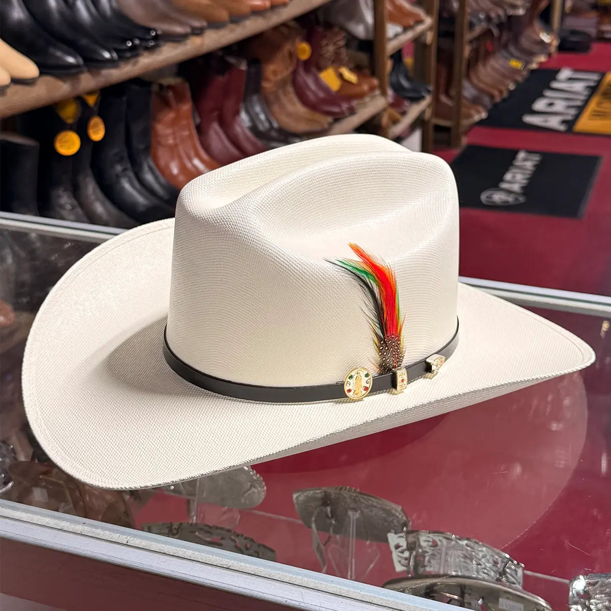 In-store display view of the Abolengo Virgen de Guadalupe 1000X Chaparral straw cowboy hat resting on a glass counter, highlighting the fine off-white weave, black hatband with gold Guadalupe pin, and colorful feather accent.