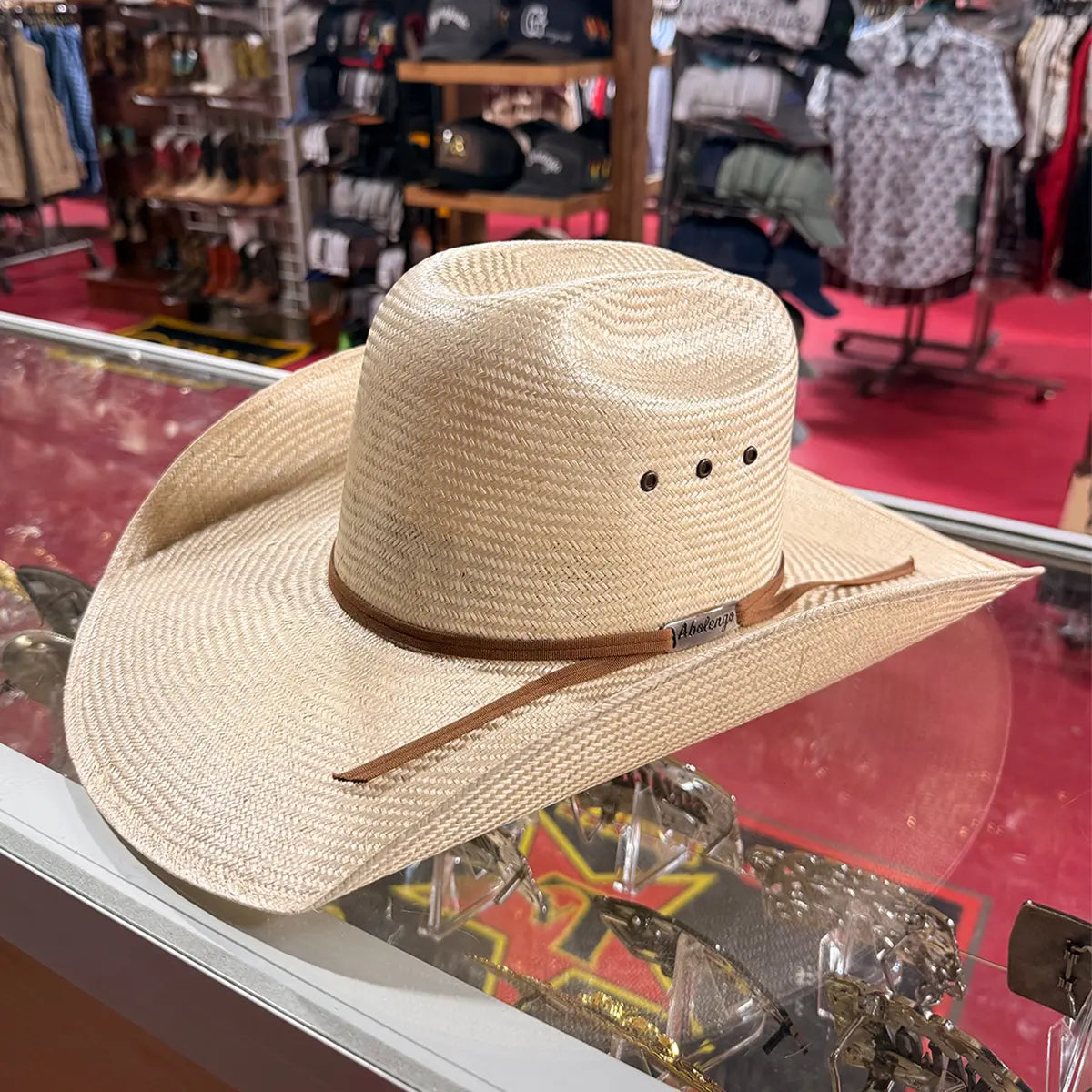 In-store display view of an Abolengo sisal straw cowboy hat resting on a glass counter, highlighting the Cattleman crown crease, side eyelet vents, brown band, and silver brand pin.