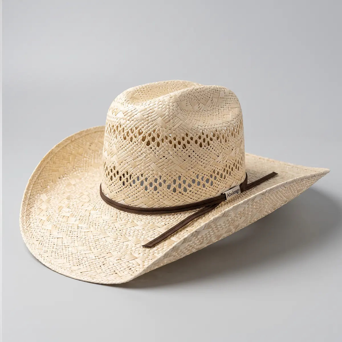 Studio three-quarter view of the Abolengo natural sisal straw cowboy hat on a neutral background showing the 5 1/4-inch vented Minnick crown and brown ribbon band.