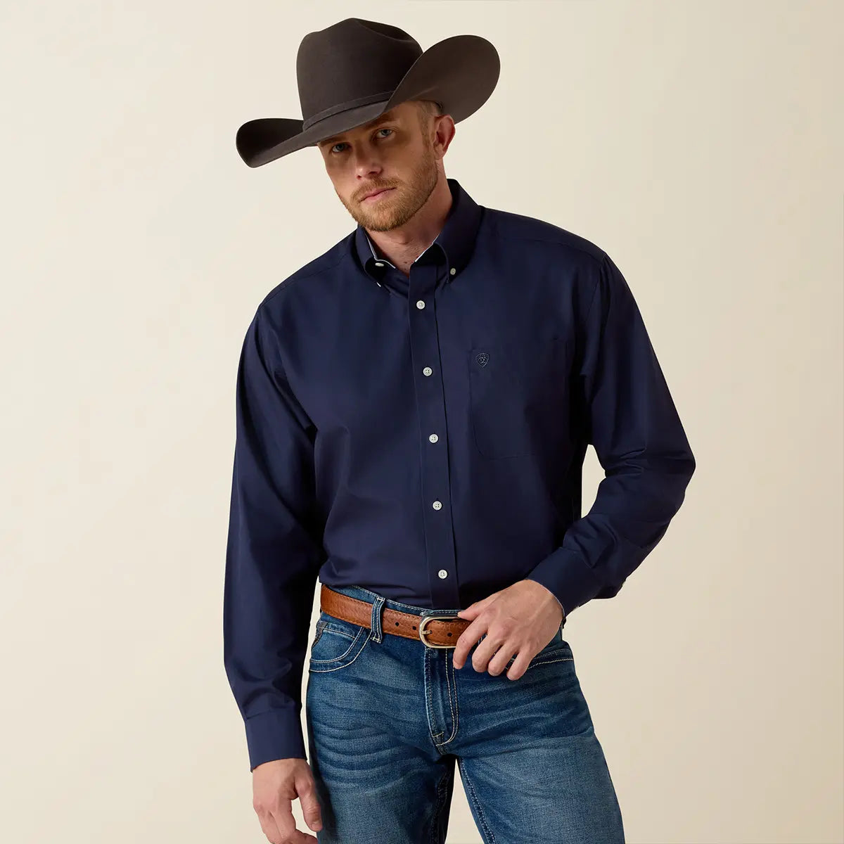 Medium front studio shot of a model wearing the Ariat Men's Navy Blue Wrinkle-Free Classic Fit shirt tucked into jeans with a dark brown cowboy hat.