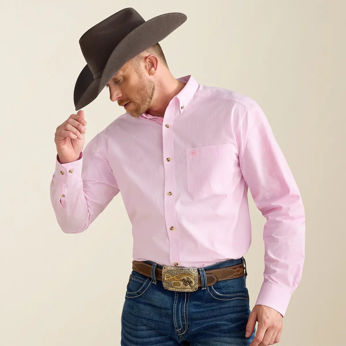 Front medium shot of a cowboy holding his dark brown hat while wearing the Ariat Men's Prism Pink Pro Series Dayne shirt tucked into jeans with a champion trophy buckle.