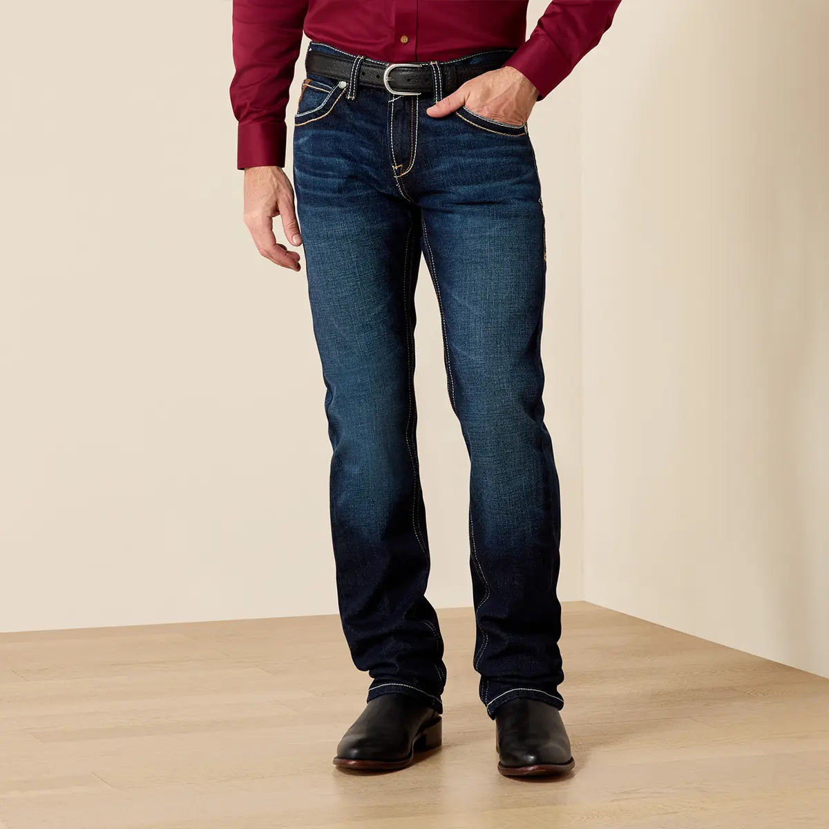Front medium shot of a model wearing Ariat Men's M7 Rocker Concord jeans in Doge Blue stretch denim with a burgundy shirt and leather belt.