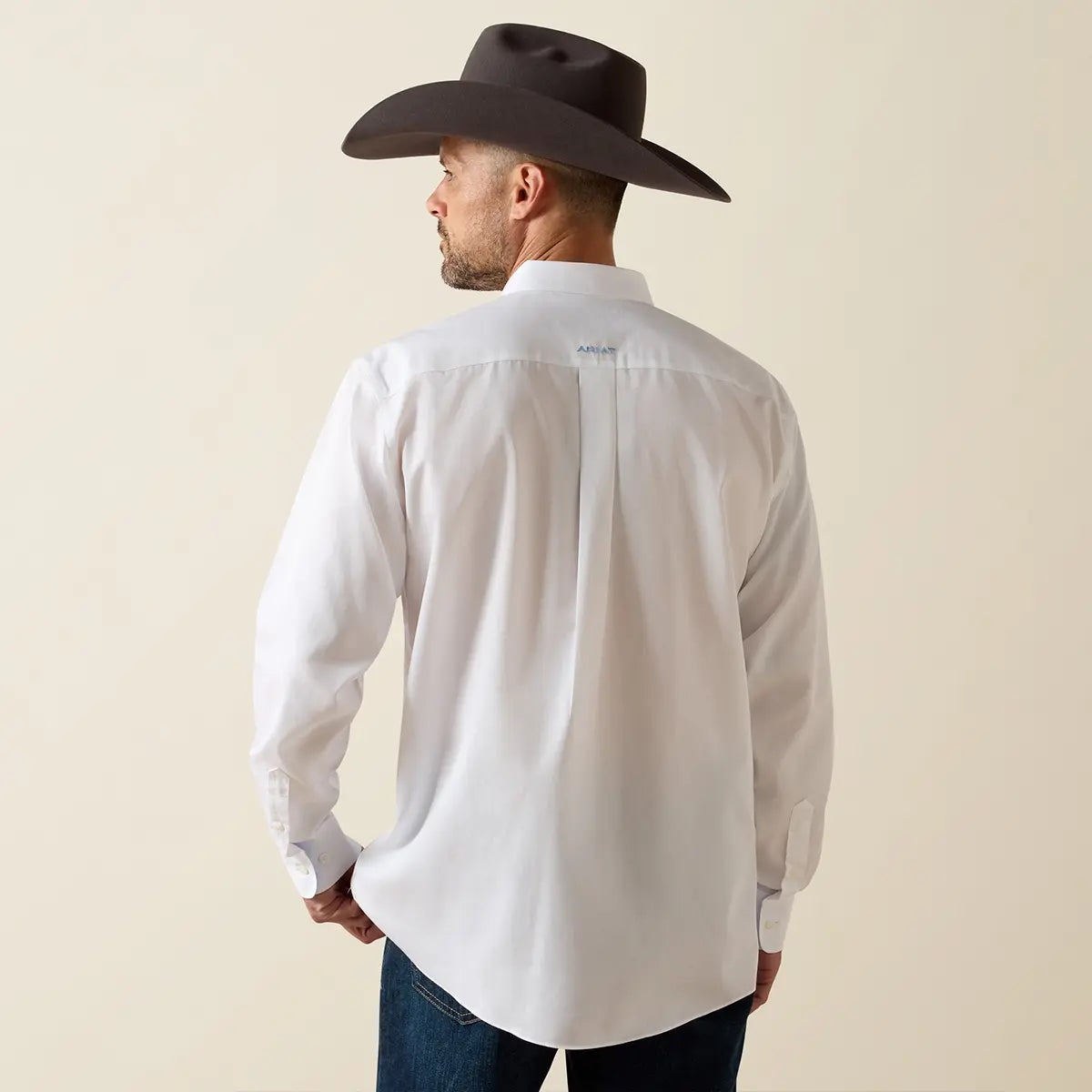 Rear view of a model wearing the Ariat white wrinkle-free classic fit shirt showing the back box pleat and blue embroidered Ariat neck logo.