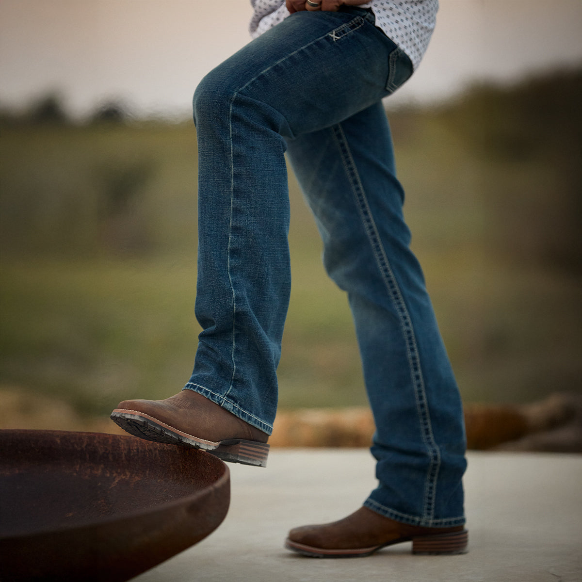 A person wearing blue denim jeans and brown leather Ariat boots, shown from the waist down in an outdoor setting with a blurred natural background.