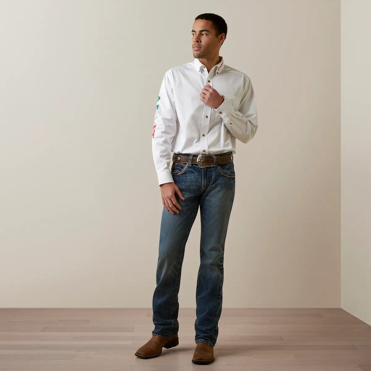 Full-length lifestyle view of a model wearing the Ariat Men's White Mexico Team Logo twill shirt tucked into blue jeans with a Western leather belt and boots.