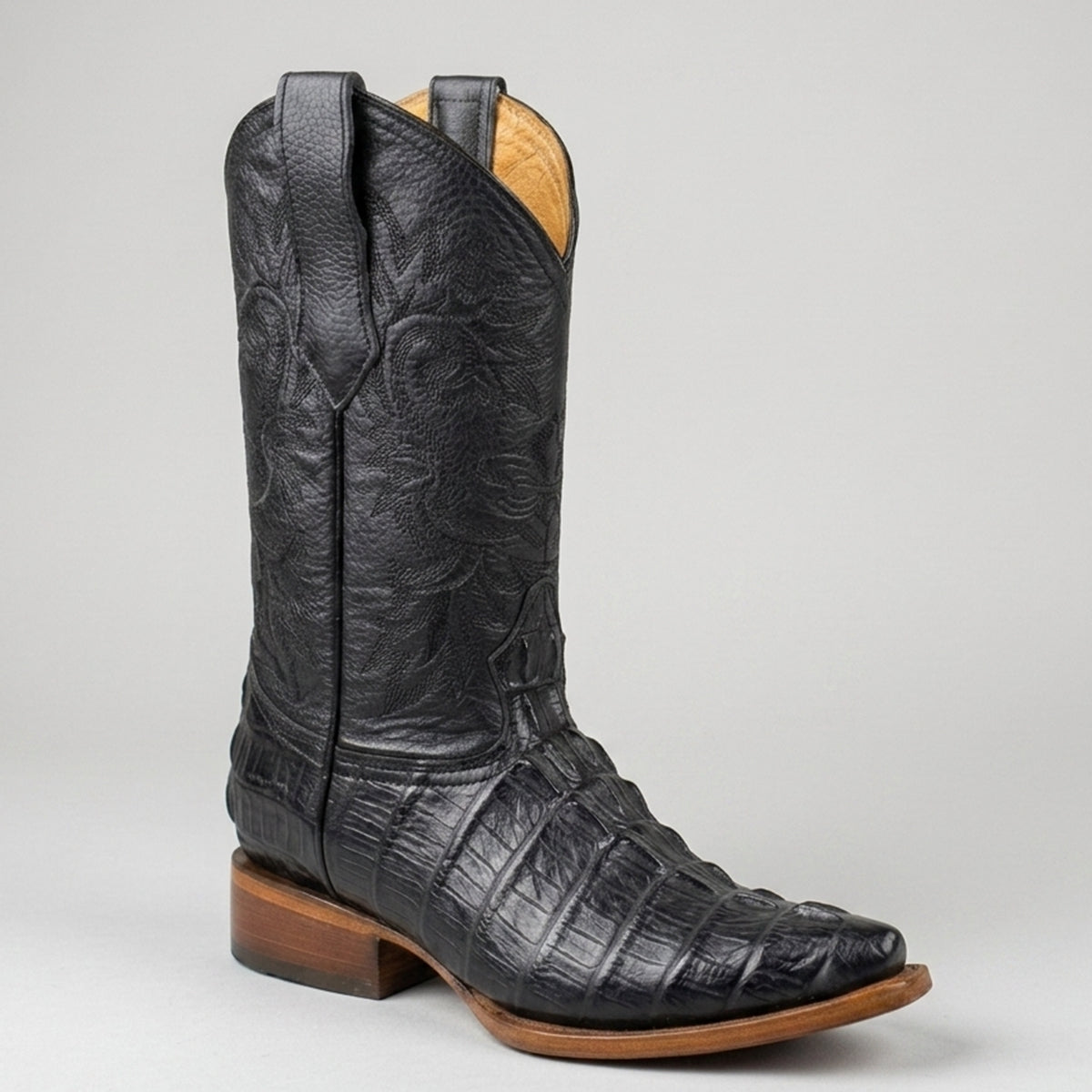 A single men's black alligator-textured leather cowboy boot with a pointed toe and stacked brown heel, shown against a neutral light gray background.