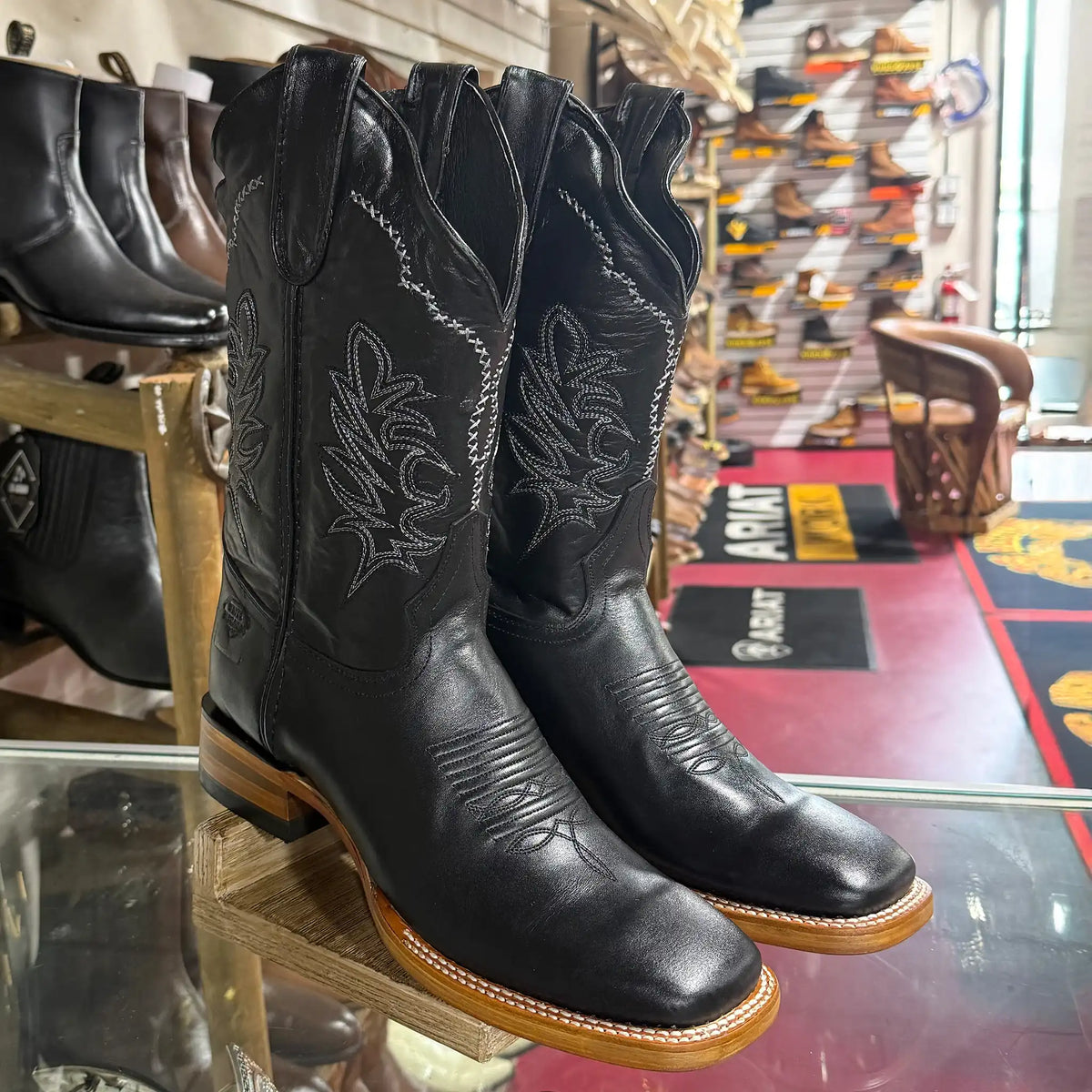 A pair of black leather western cowboy boots with white embroidered floral detailing, displayed on a wooden stand inside a store.