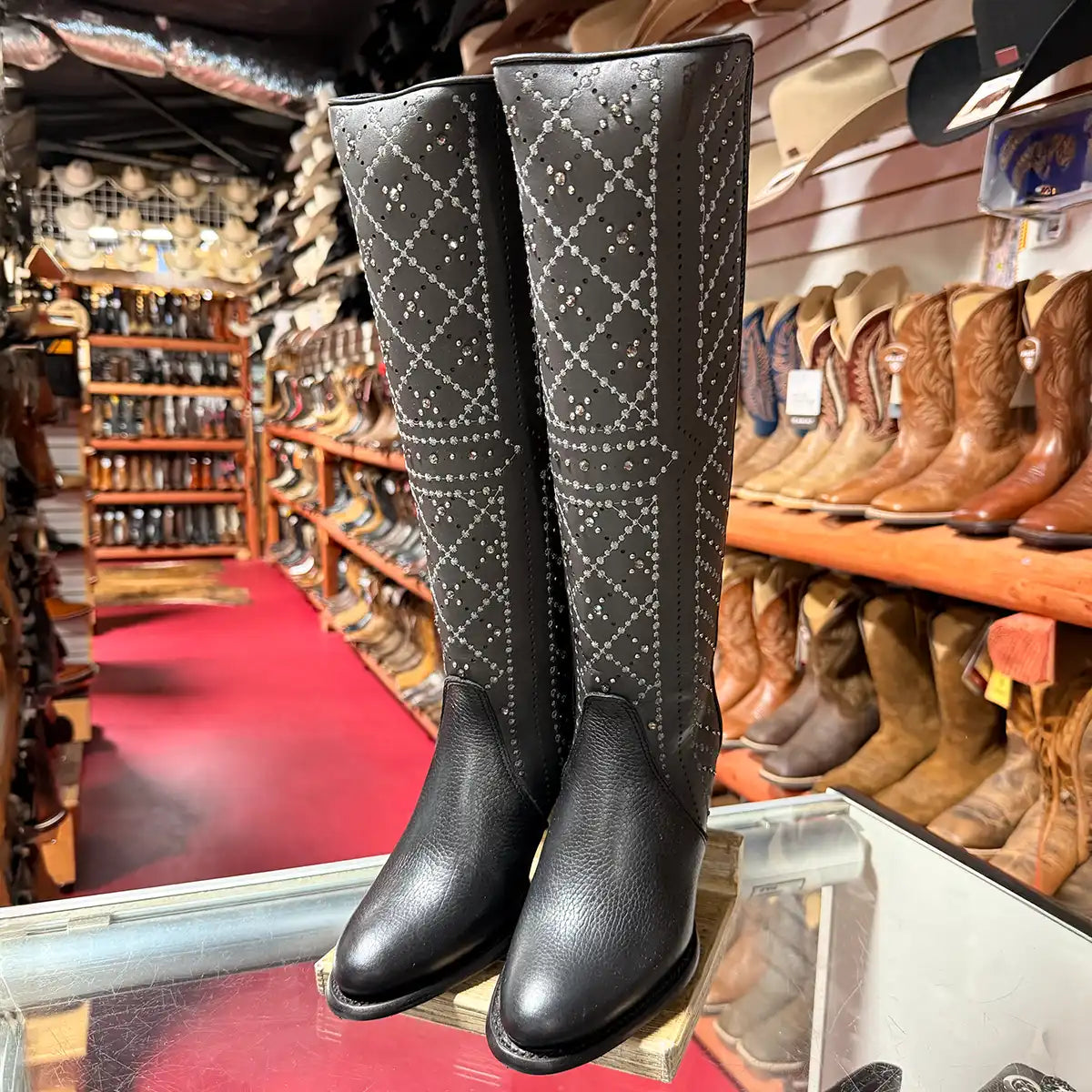 A pair of Cuadra women's black knee-high western boots with silver rhinestone studded shafts, displayed on a glass counter inside a boot shop with shelves of other boots in the background.