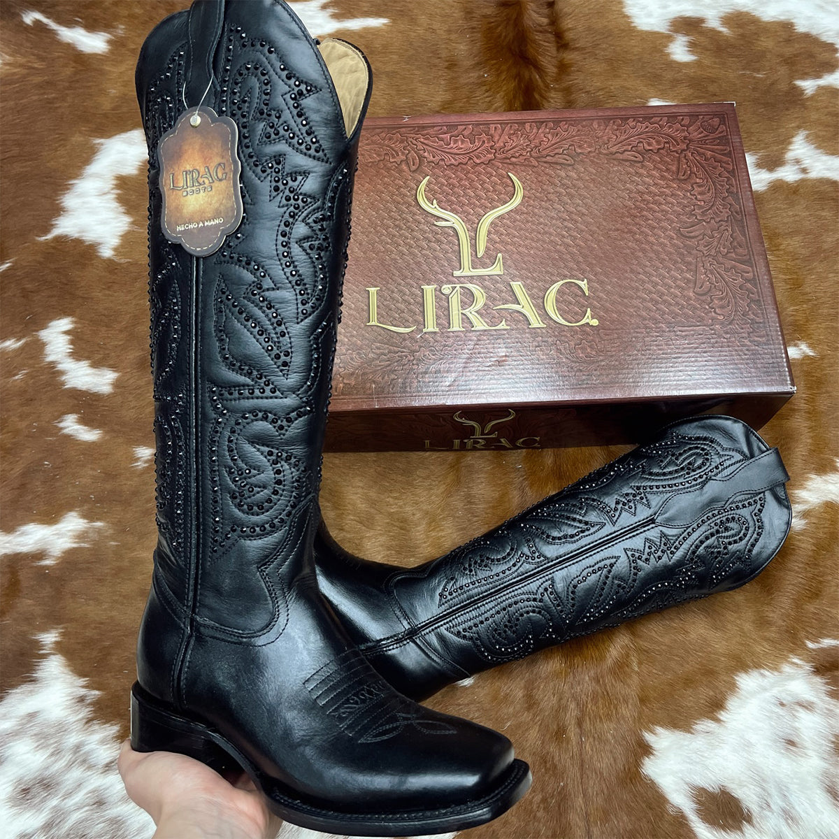 A pair of women's Lirac black leather square toe cowgirl boots with intricate western embroidery, shown with their brown branded box on a brown and white cowhide rug.