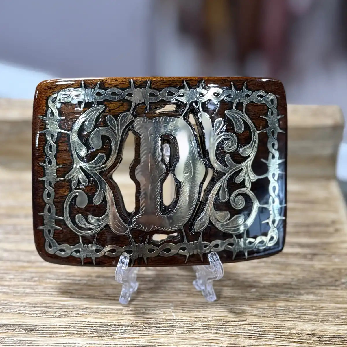 Front view of the handcrafted mesquite wood belt buckle showing the metal Monogram D inlay and barbed wire frame displayed on an acrylic stand.