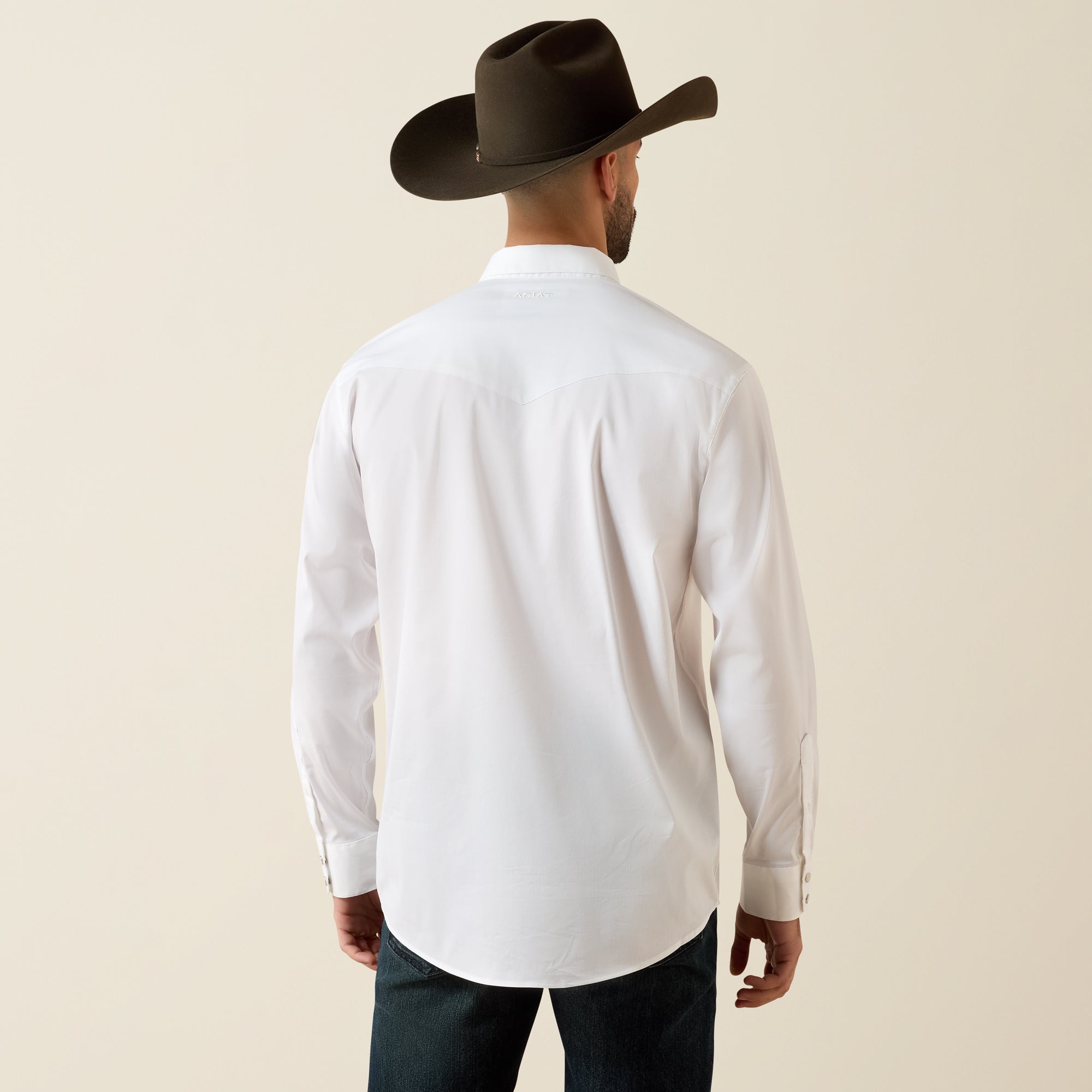 A man viewed from behind wearing a white long-sleeve button-down shirt, dark jeans, and a dark brown cowboy hat against a solid light beige background.