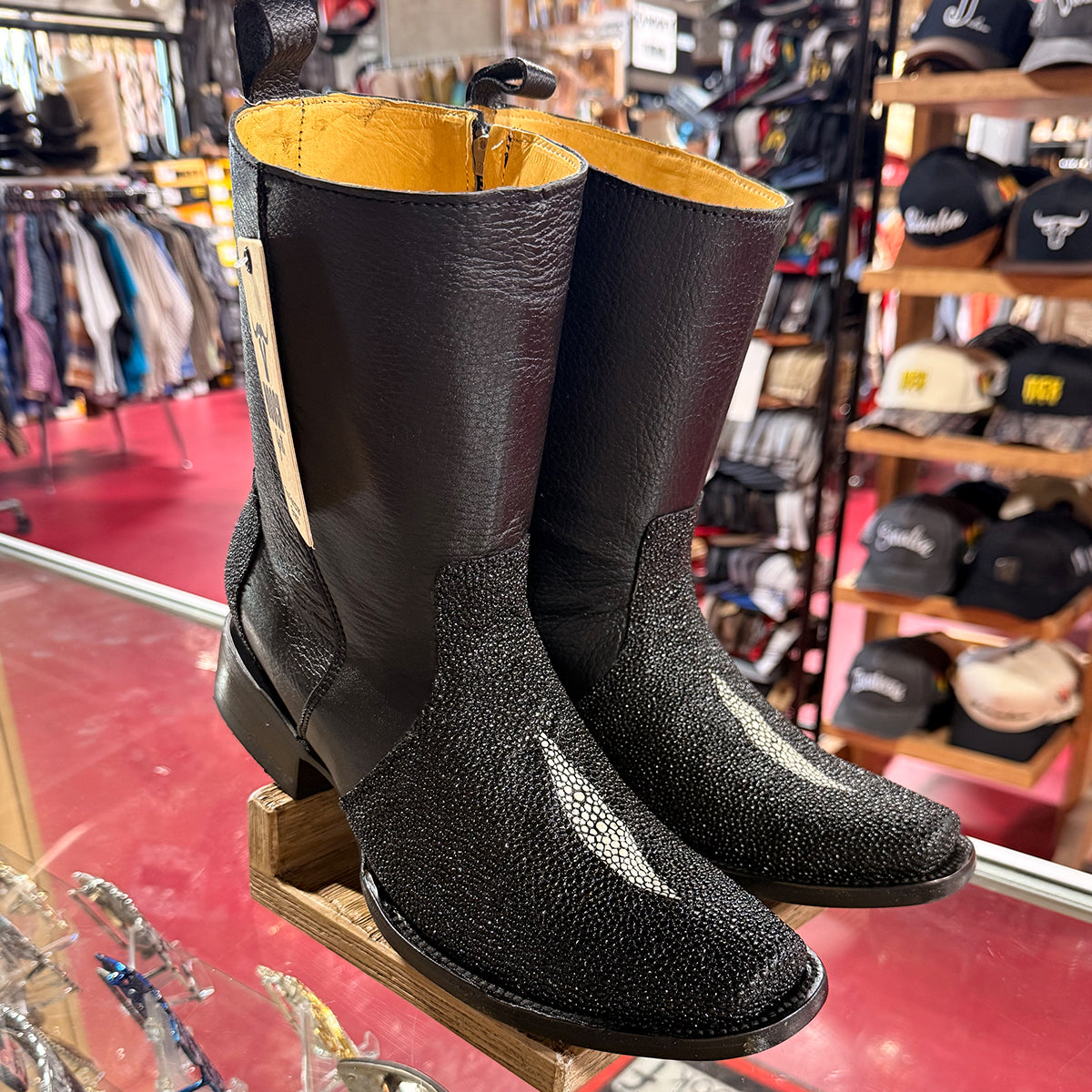 A pair of men's black stingray leather print cowboy boots, displayed on a wooden stand inside a western store.