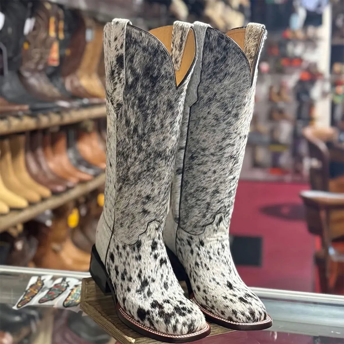 Front angled view of black and white cow hair-on hide women's square toe cowboy boots displayed on a wooden stand inside a retail western apparel store.