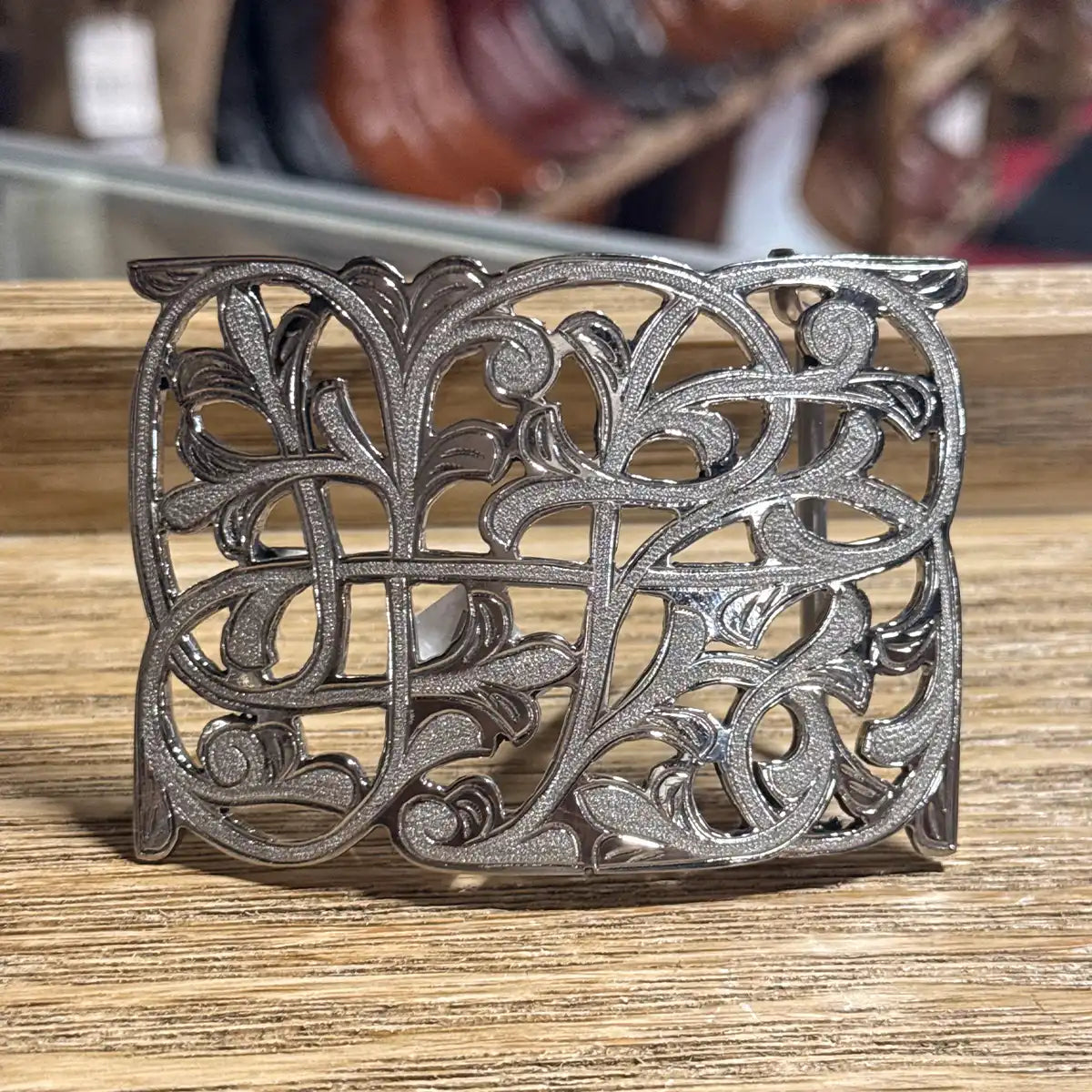 Counter display view of the stainless steel openwork belt buckle with floral filigree scrollwork on a wooden display.