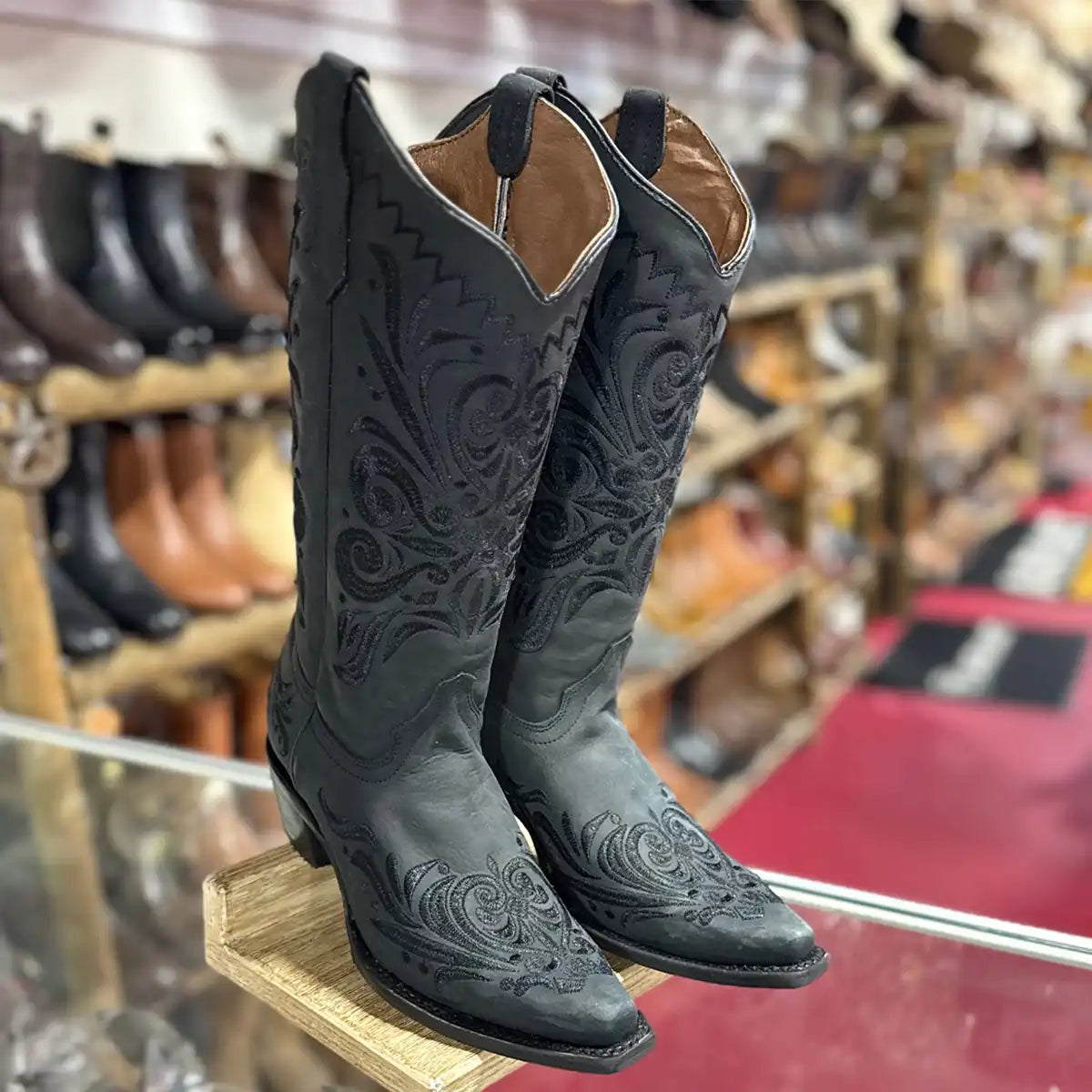 Corral women's black leather cowboy boots with intricate embossed western stitching, displayed on a wooden stand inside a western store.