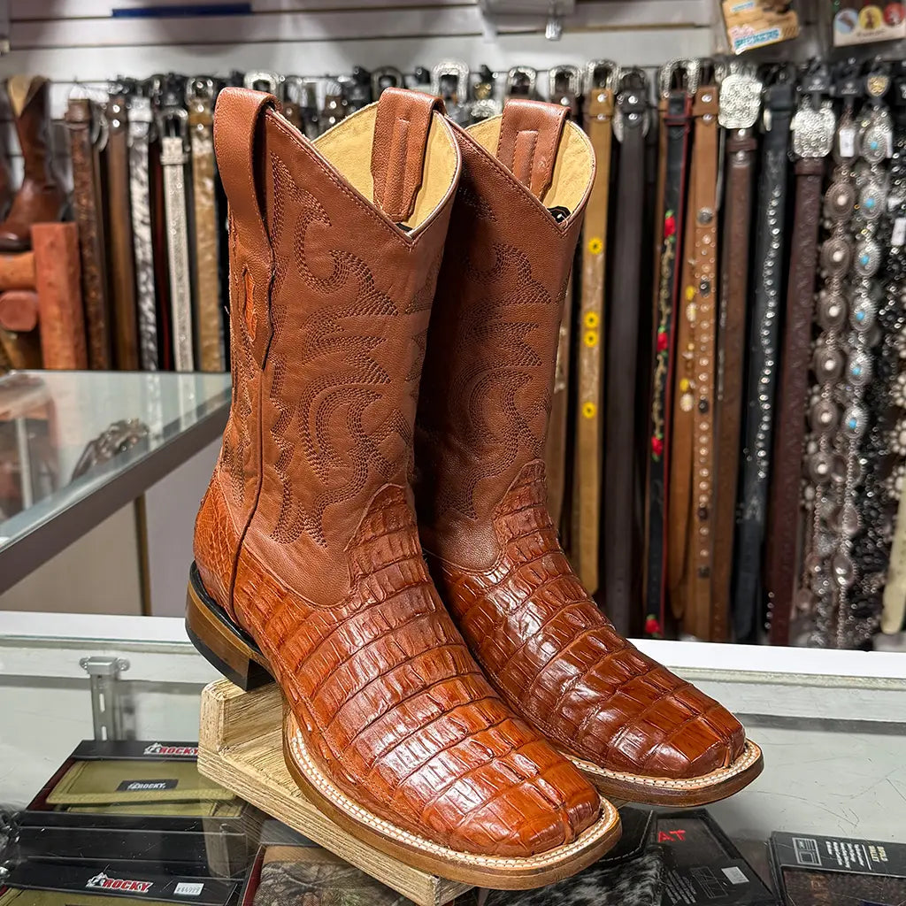 Retail display view of the Los Altos cognac caiman tail boots sitting on a glass counter inside a Western storefront surrounded by leather belts.