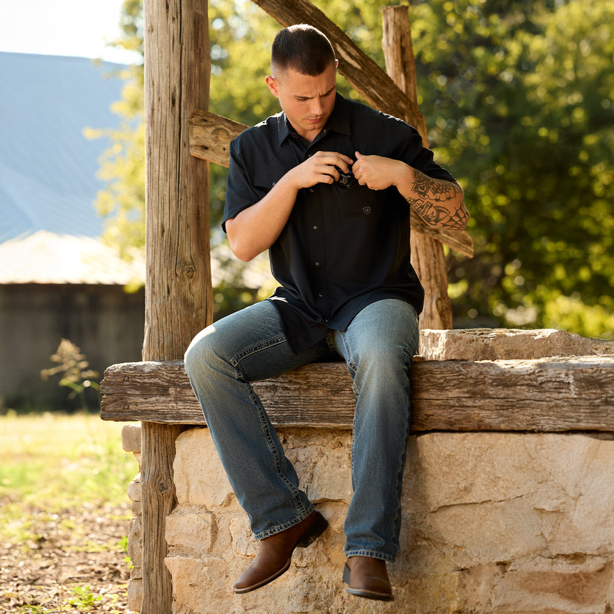 A man wearing a black short-sleeve button-up shirt and blue jeans with Ariat boots siting on a rustic stone and wood bench outdoors, adjusting the collar of his shirt.