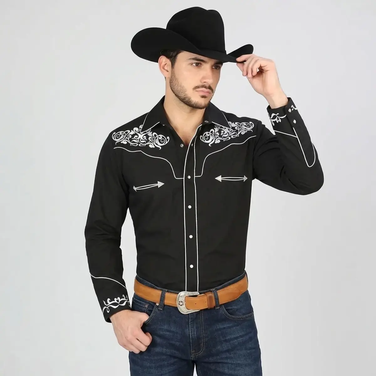 Front view of a male model wearing a men's black Western snap shirt featuring white scrollwork embroidery on the shoulders, white piping on the yokes, white arrow smile accents, and pearl snap buttons with a black cowboy hat and jeans.