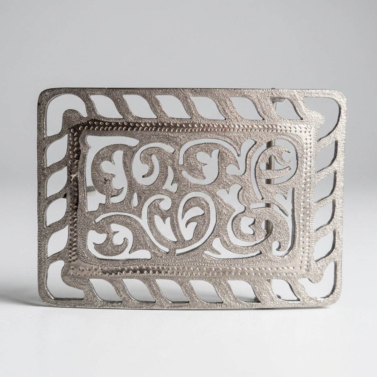 An ornate silver metal belt buckle with intricate filigree cutouts and decorative scrollwork, shown on a neutral light gray background.