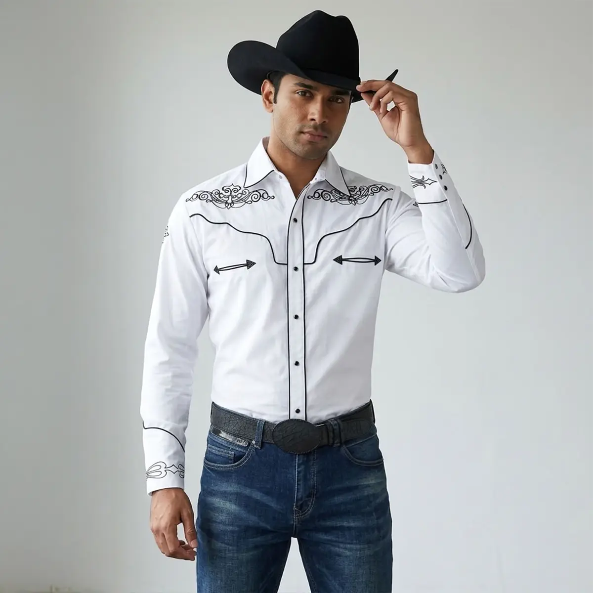 Front view of a male model wearing a men's white long-sleeve Western snap shirt with black scroll embroidery on the yokes, black contrast piping along the placket, smiley arrow accents, black cowboy hat, and jeans.