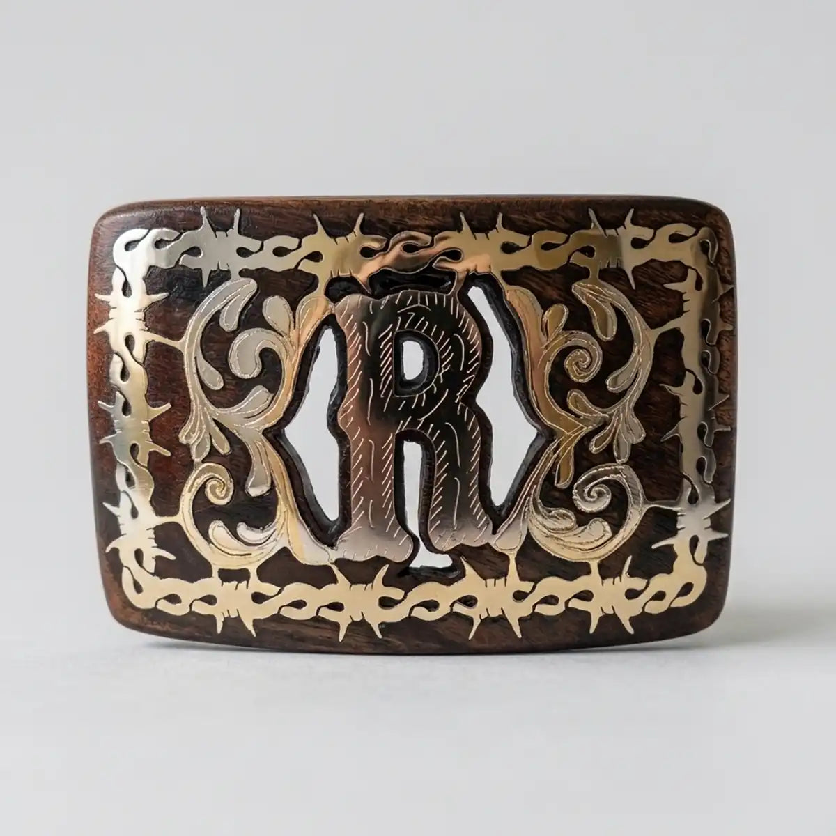 Handcrafted mesquite wood Western belt buckle with polished metal Monogram R inlay, floral scrollwork, and barbed wire border frame on light grey background.