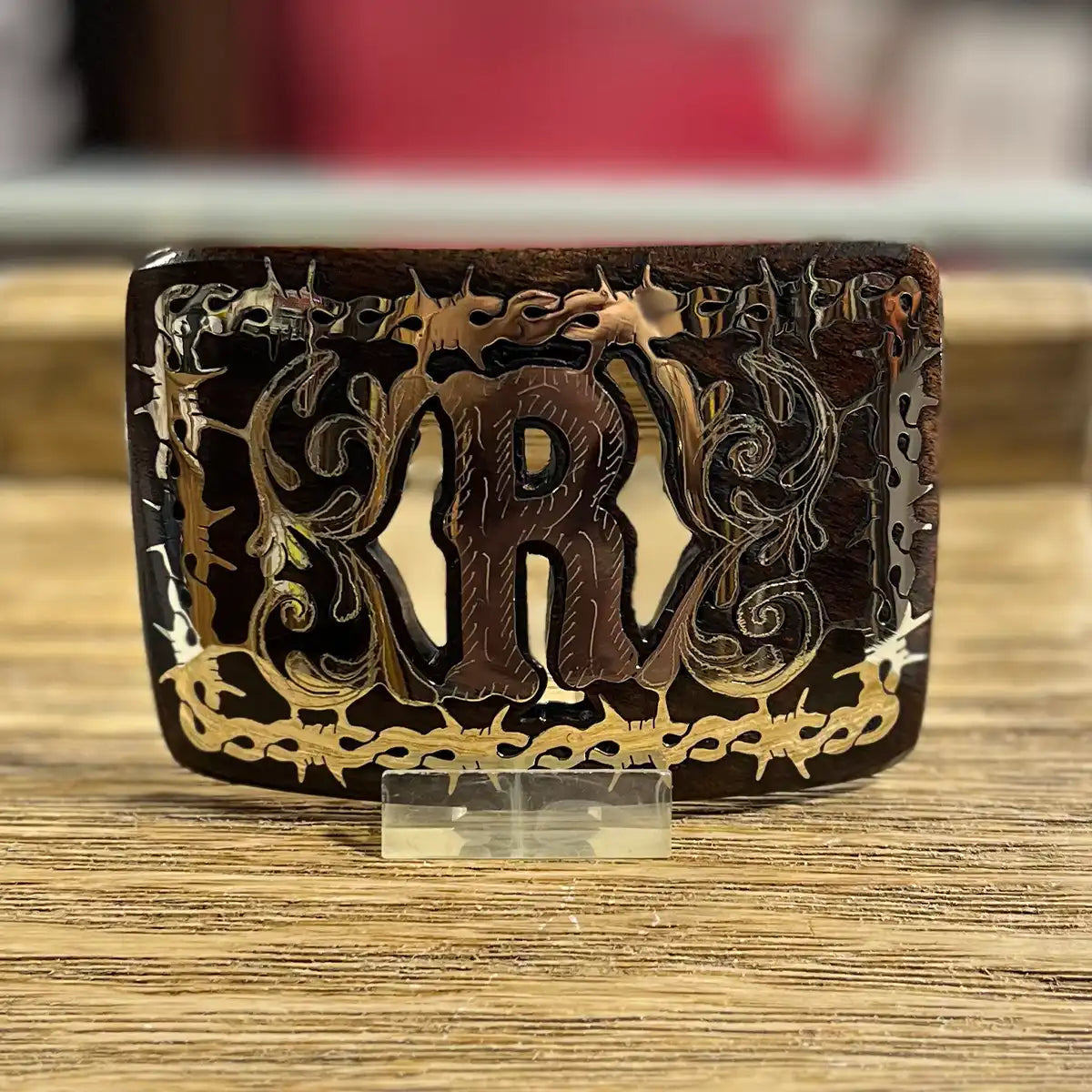 Handcrafted mesquite wood Western belt buckle with polished metal Monogram R inlay, floral scrollwork, and barbed wire border frame.