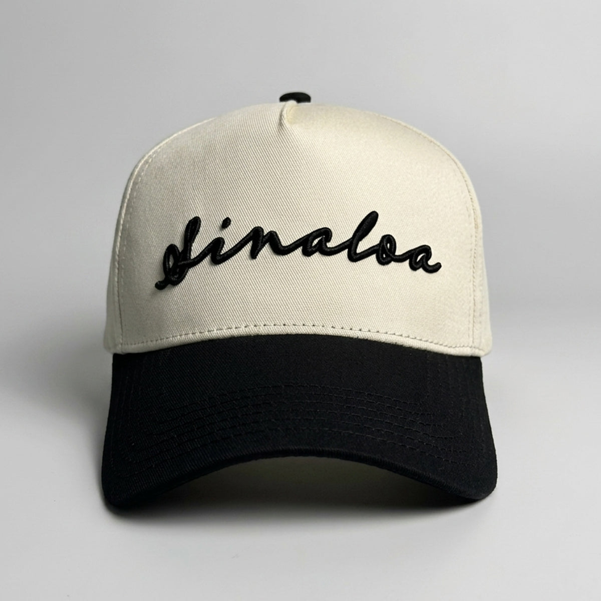 A two-tone baseball cap with an off-white crown and a black curved brim, featuring the word 'Sinaloa' embroidered in black cursive script across the front.