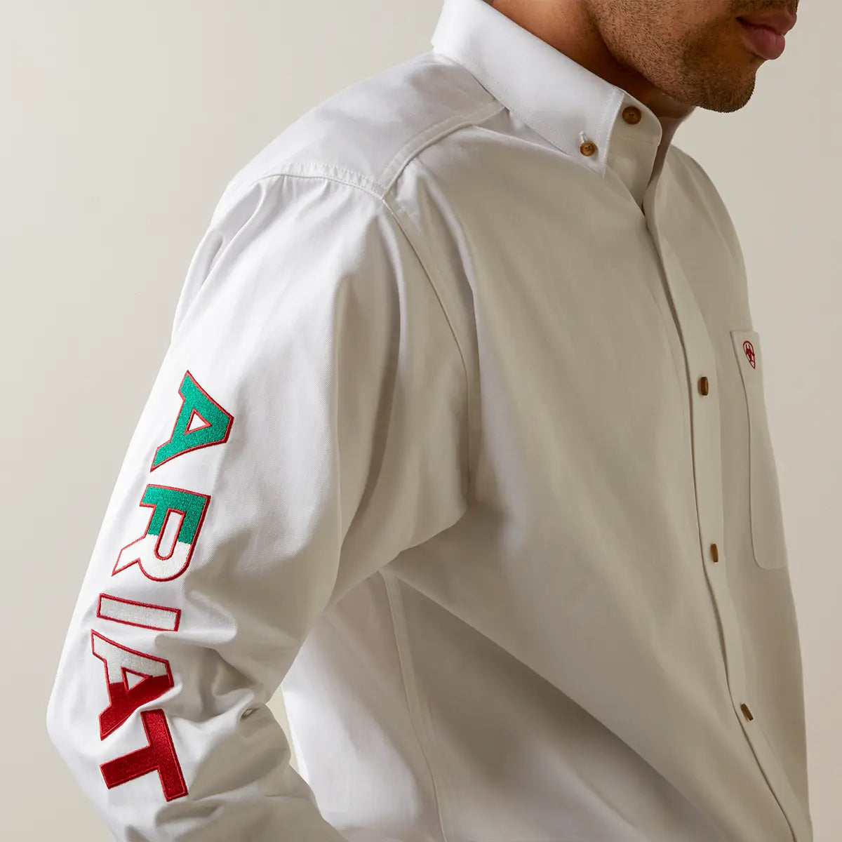 Close-up view of the right sleeve highlighting the detailed red, green, and white embroidered ARIAT block lettering on white cotton twill.