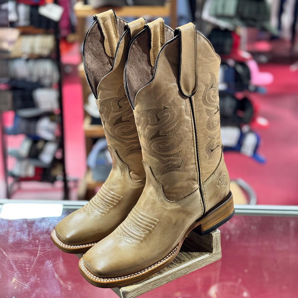 Pair of women's tan cowboy boots on a glass display stand with a store interior background.