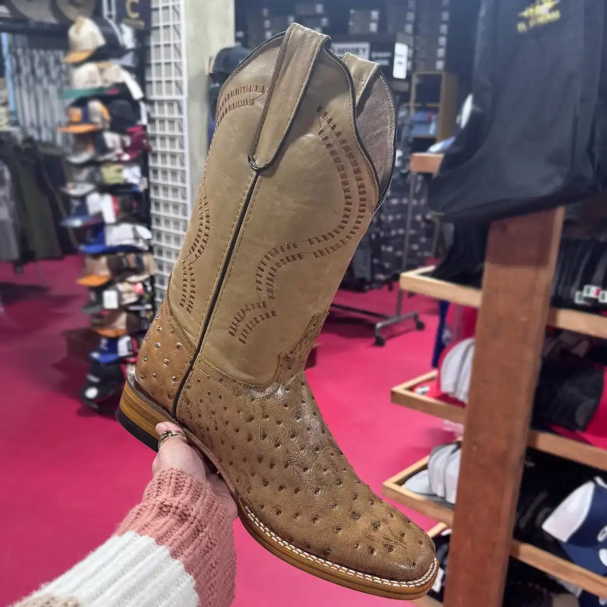 Brown ostrich print square toe cowboy boot held in a store setting with shelves and products in the background.