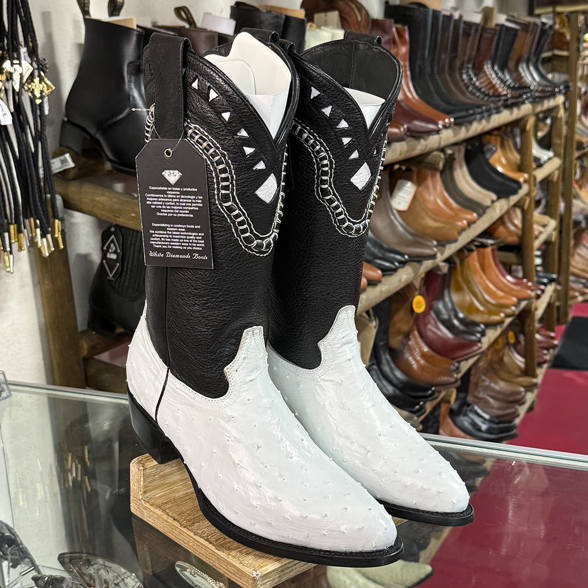 A pair of men's ostrich western cowboy boots displayed on a wooden stand inside a shoe store. The boots feature a black upper with white stitched detailing and a white lower ostrich vamp with an embossed texture.