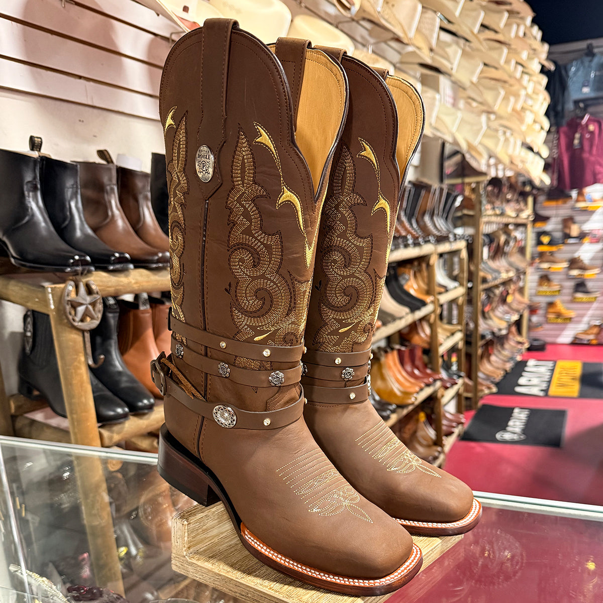 A pair of women's brown western cowboy boots with gold embroidery, silver conchos, and decorative straps, displayed on a wooden stand inside a western store.