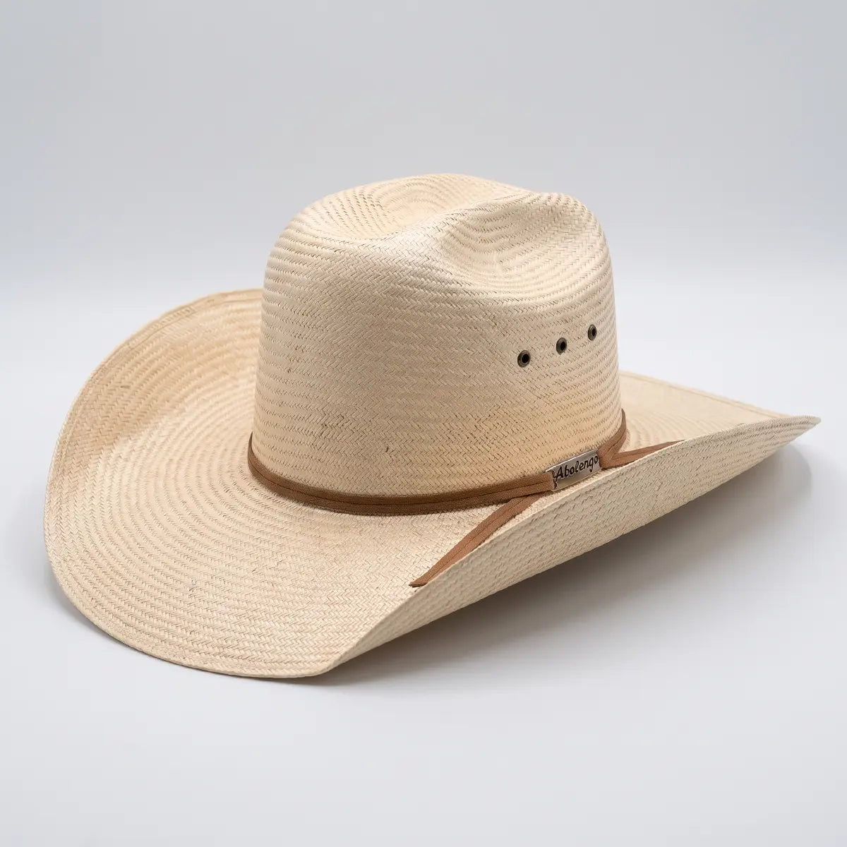 Studio three-quarter view of the Abolengo sisal straw cowboy hat on a white background, highlighting the curved brim, Cattleman crown, side ventilation eyelets, brown band, and silver logo pin.