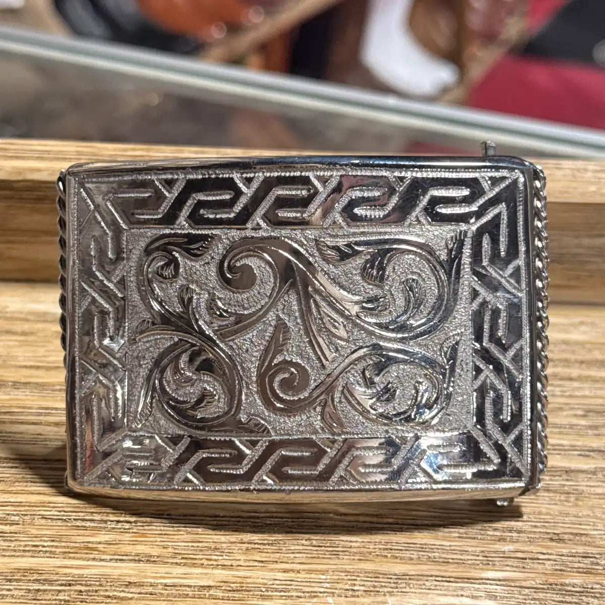 In-store display view of the 3.5" x 2.5" stainless steel floral engraved belt buckle resting on a wooden counter top.