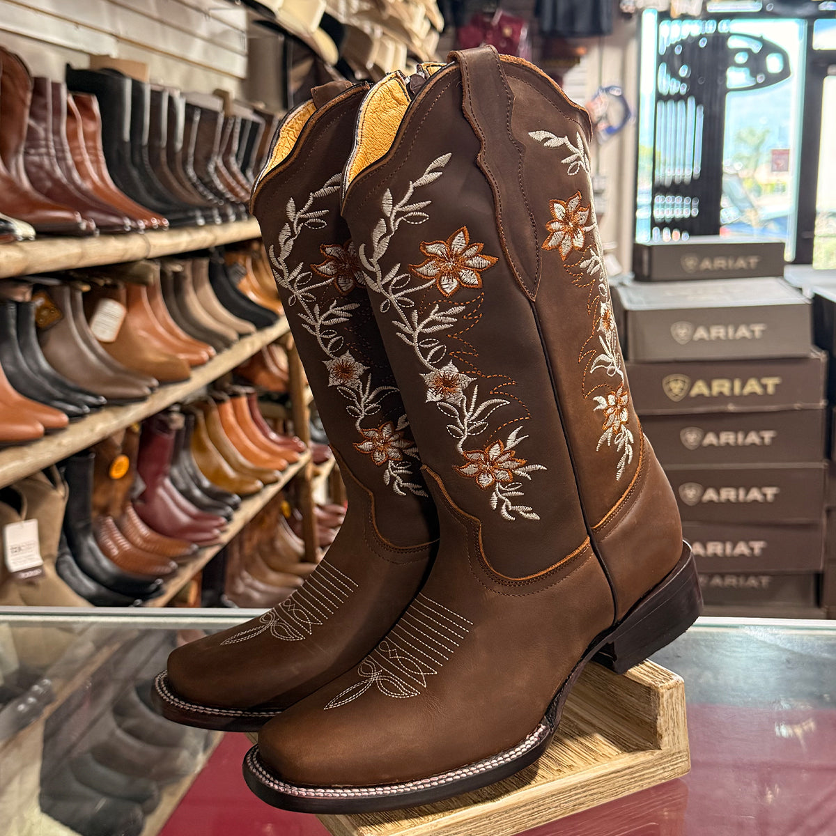 La Sierra brown leather square toe cowgirl boots with floral embroidery, displayed on a wooden stand inside a western wear store.
