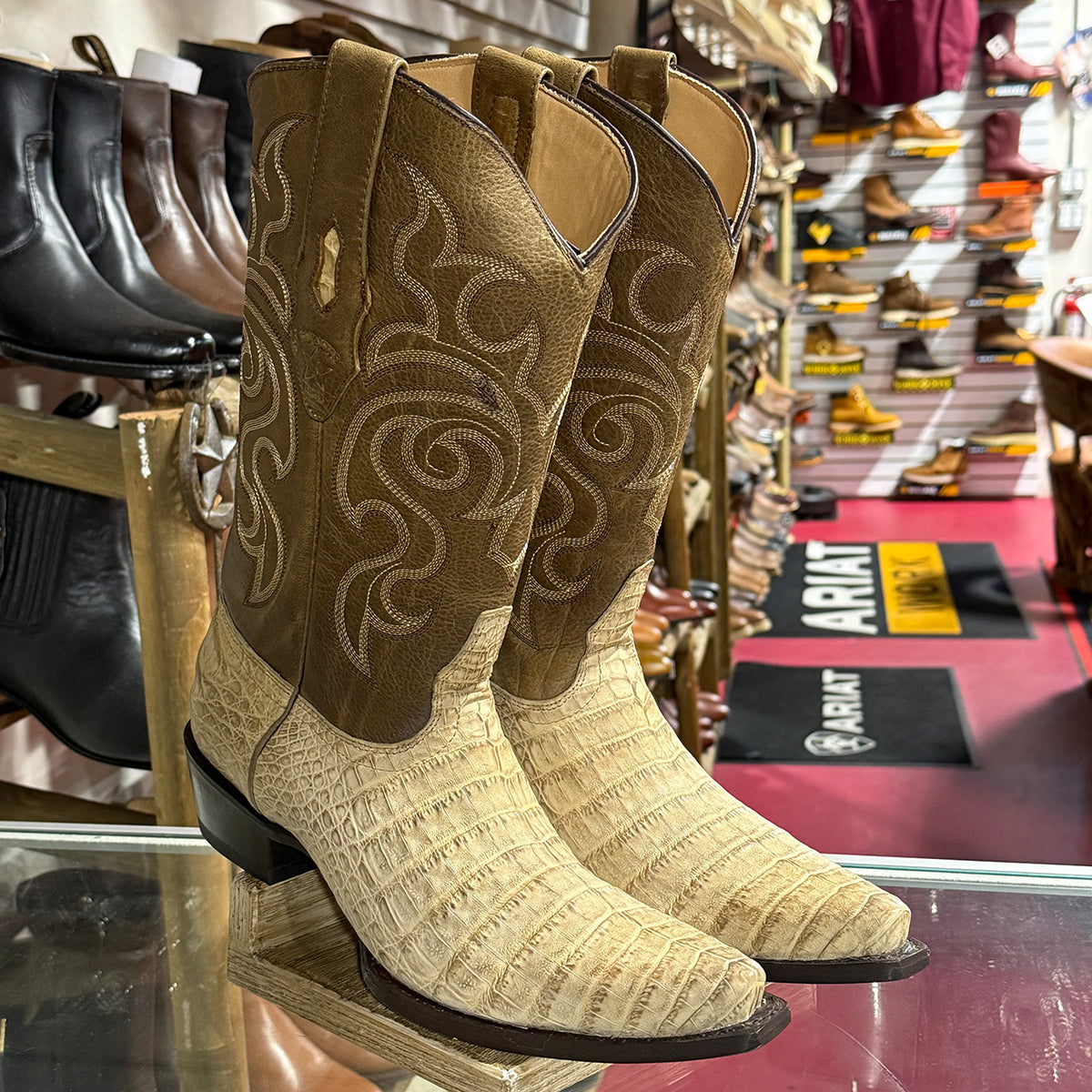 A pair of cowboy boots displayed on a glass counter inside a shoe store. The boots feature a tan leather shaft with white embroidered scrollwork and genuine beige caiman belly foot.