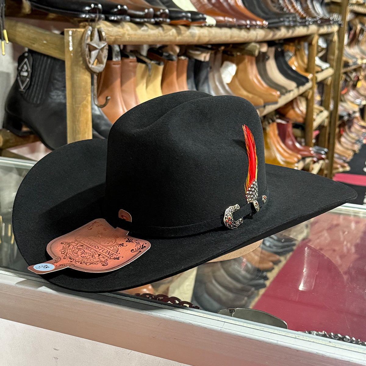 Tombstone 50x Chaparral black beaver felt cowboy hat with a red and orange feather, a silver concho-style ornament, and a tooled brown leather brand patch, displayed on a glass case in a western store background.
