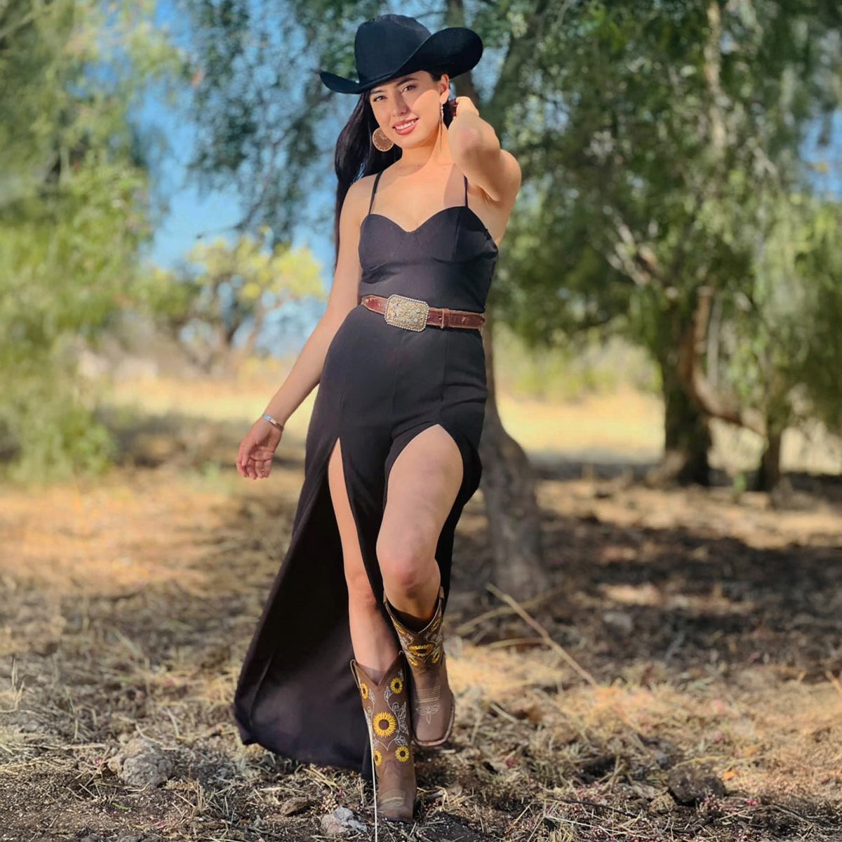 A woman stands outdoors in a wooded area wearing a black cowboy hat, a black spaghetti-strap maxi dress with a thigh-high slit and a brown belt, and brown sunflower cowboy boots.