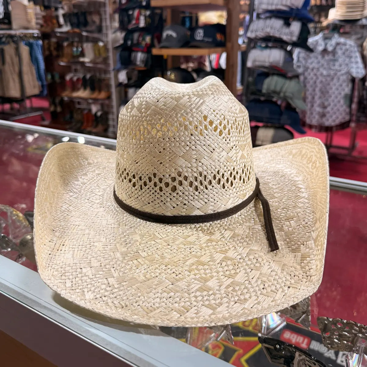 In-store display view of the Abolengo natural sisal straw cowboy hat resting on a glass counter, highlighting the symmetrical curved brim, all-around open-weave ventilated Minnick crown, and chocolate brown ribbon hatband.