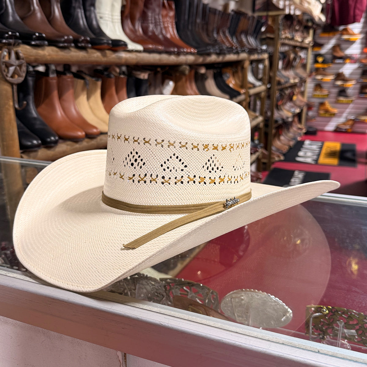 A light beige woven Tombstone cowboy hat with a tan band and embroidered details, displayed on a glass counter inside a western store.